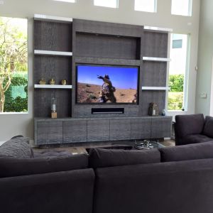 A grey oak media wall with white floating shelves and a large television, in a bright double-height living room with a corner sofa.