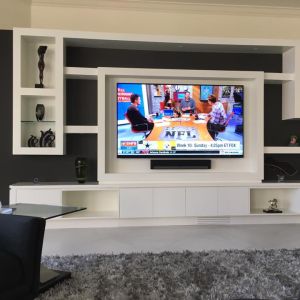 A white built-in wall unit with a dark inset panel framing a large television showing a live American football broadcast, with a soundbar below.