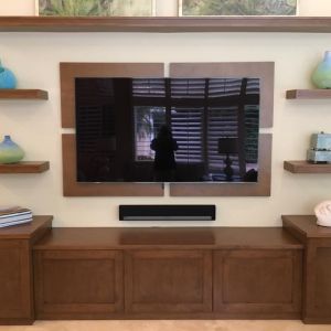 A television mounted on a walnut panel between dark wood floating shelves, above a long low cabinet, in a cream-walled living room.
