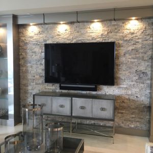 A television mounted on a stacked stone wall lit by downlights, above a mirrored sideboard set with glassware.