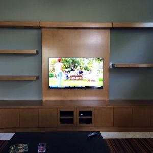A maple media wall with floating shelves and a wall-mounted television, above a cabinet housing audio-visual equipment.