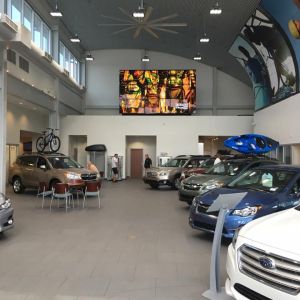 A large video screen mounted high on the wall of a car dealership showroom, above rows of cars and display racks.