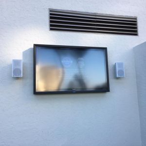 An outdoor television in a weatherproof enclosure mounted on a white stucco patio wall, with a matching outdoor speaker either side.