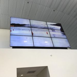 A nine-screen video wall mounted high in a commercial lobby with a corrugated metal ceiling.