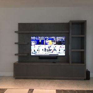 A grey oak media wall with open shelving either side of a large television showing an American football game, with a soundbar mounted beneath it.