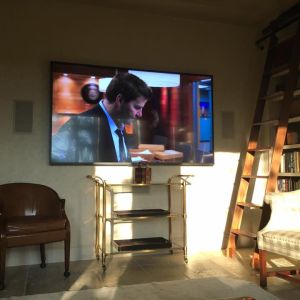 A television wall-mounted above a brass and glass console table, beside a wooden library ladder and a leather armchair.