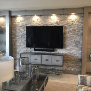 A large flat-screen television wall-mounted on a stacked stone feature wall, lit by four downlights, above a mirrored console table in a living room.