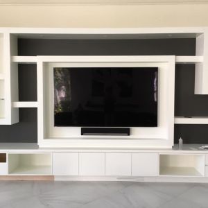 A white built-in wall unit with dark grey recesses, framing a large wall-mounted television and soundbar above a marble floor.