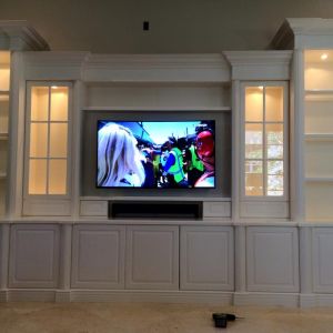 White built-in cabinetry with illuminated glass-fronted display cabinets either side of a wall-mounted television and soundbar.