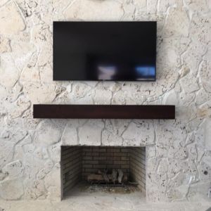 A television wall-mounted above a dark wood mantel beam on a rough limestone fireplace wall with an open firebox below.